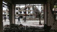 A picture taken on February 17, 2018 shows a man walking next to another pushing a bicycle loaded with a carpet, down a rain-soaked street past damaged and destroyed buildings in the Syrian rebel-held enclave of Arbin in the Eastern Ghouta near the capita