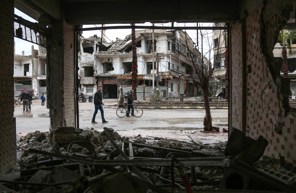 A picture taken on February 17, 2018 shows a man walking next to another pushing a bicycle loaded with a carpet, down a rain-soaked street past damaged and destroyed buildings in the Syrian rebel-held enclave of Arbin in the Eastern Ghouta near the capita