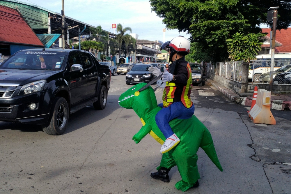 Thai police sargent Tanit Bussabong directs traffic wearing a costume where he appears to ride a T-rex dinosaur outside a school in Nakhon Nayok on June 4, 2018. AFP / Stephen J. BOITANO
 