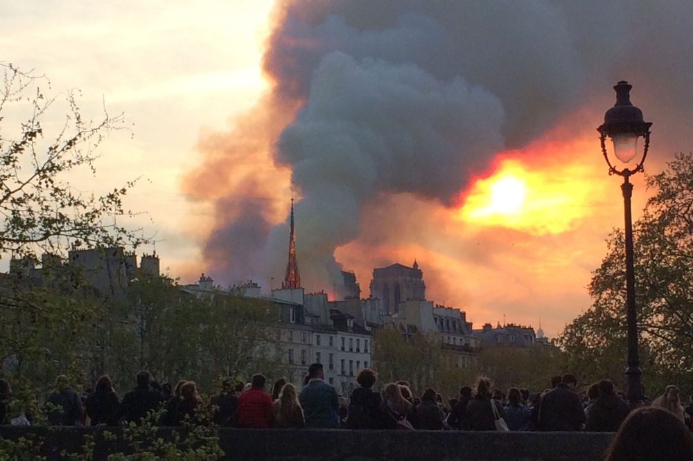 A picture taken with a mobile phone shows people looking at smoke and flames rising during a fire at the landmark Notre-Dame Cathedral in central Paris on April 15, 2019, potentially involving renovation works being carried out at the site, the fire servi