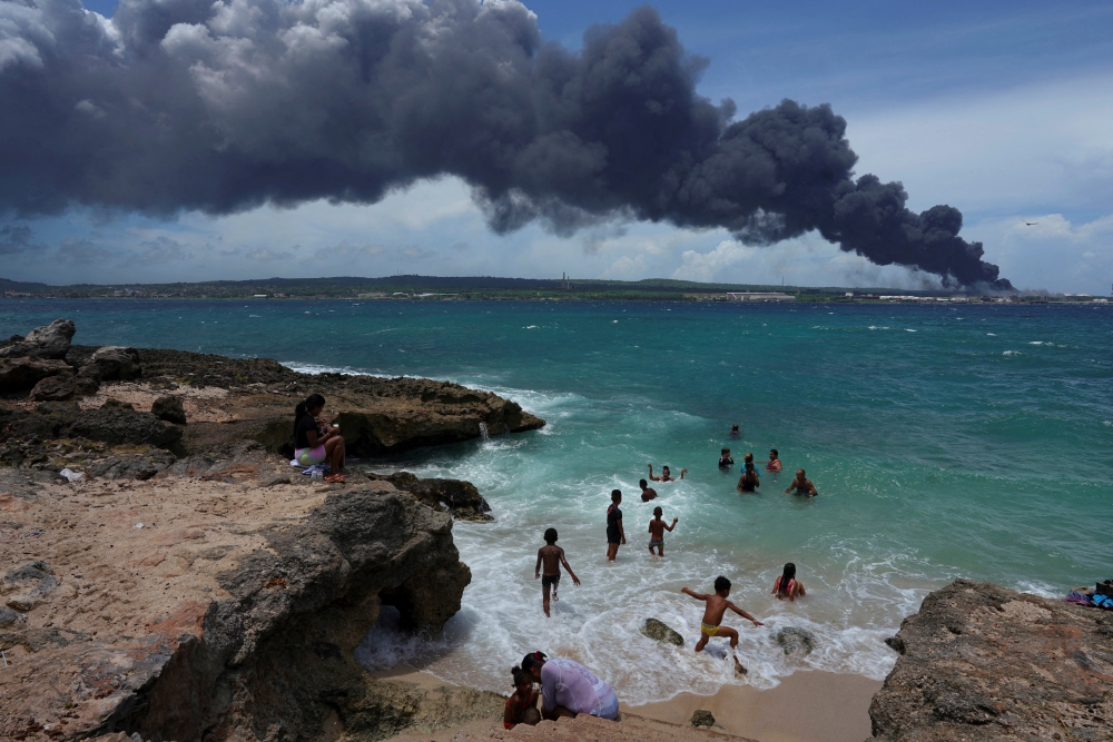 People enjoy the beach near the smoke of a fire over fuel storage tanks that exploded near Cuba's supertanker port in Matanzas, Cuba, August 6, 2022. REUTERS/Alexandre Meneghini