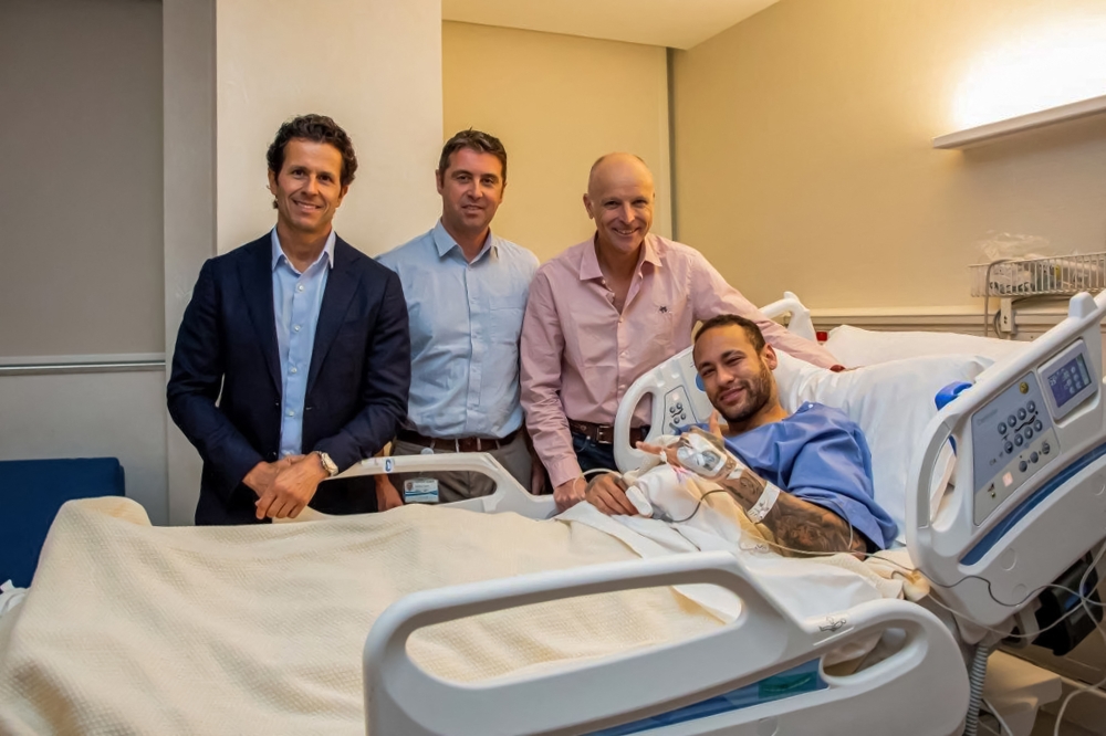 This handout picture provided by Aspetar Hospital in Doha on March 10, 2023 shows Paris Saint-Germain's Brazilian forward Neymar da Silva Santos Junior (R) gesturing in bed after after undergoing surgery at the facility, accompanied by the hospital's Chief Medical Officer Dr Pieter D'Hooghe (2nd-L), ankle surgeon Dr Pierre James Calder (2nd-R), and Brazilian surgeon and national team doctor Rodrigo Lasmar (L). (Photo by Aspetar Hospital Doha / AFP)