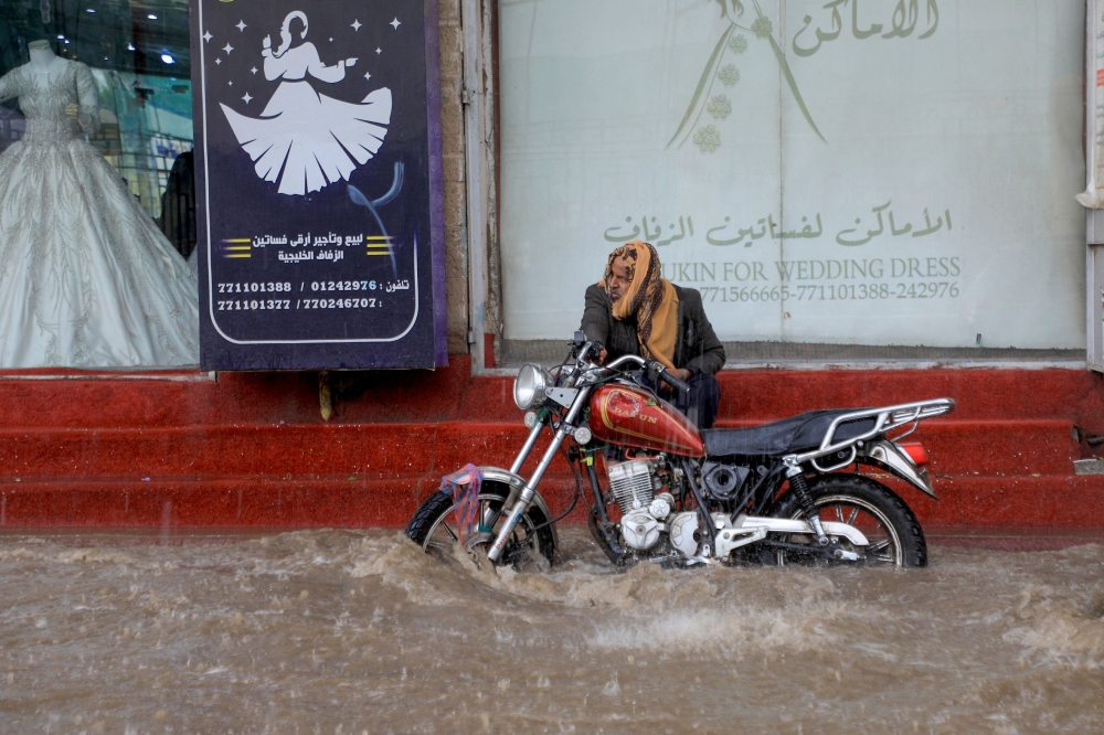 A man sits by his motorcycle on a shop's stairs as water floods a street amid heavy rainfall in Sanaa, Yemen, on March 31, 2023. (Photo by Mohammed Huwais / AFP)