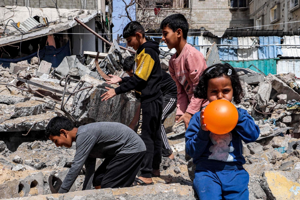 A girl blows a balloon as behind her boys search through the rubble of a destroyed building in Rafah in the southern Gaza Strip on April 5, 2024. (Photo by Mohammed Abed / AFP)
 