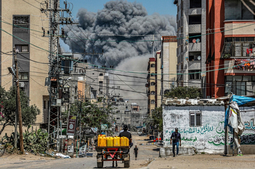 A cloud of smoke erupts down the road from an explosion as a man drives an animal-drawn cart loaded with jerrycans in Nuseirat in the central Gaza Strip on April 17, 2024. (Photo by AFP)