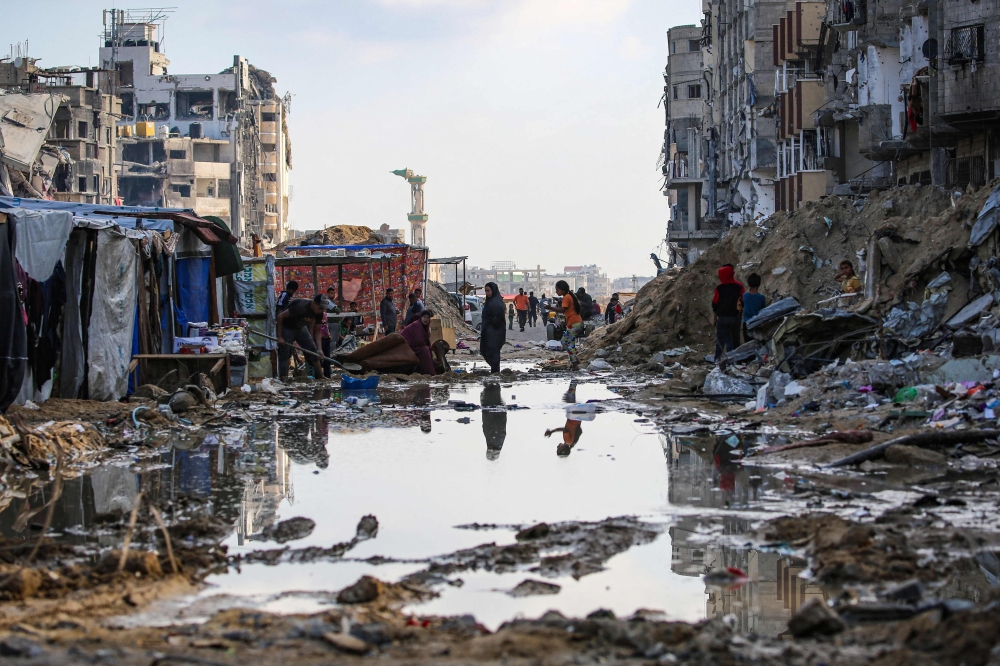 Displaced Palestinians walk around a puddle in front of destroyed buildings and tents in Khan Yunis in the southern Gaza Strip on May 16, 2024. (Photo by AFP)