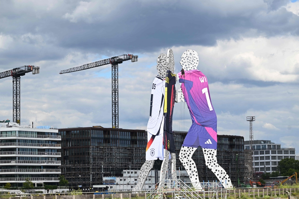 A worker climbs the 'Molecule Man' sculpture, designed by US artist Jonathan Borofsky wearing the three different Germany's national kits in the water of the river Spree in Berlin on June 13, 2024, ahead of the UEFA Euro 2024 European Football Championship. (Photo by Ralf Hirschberger / AFP)