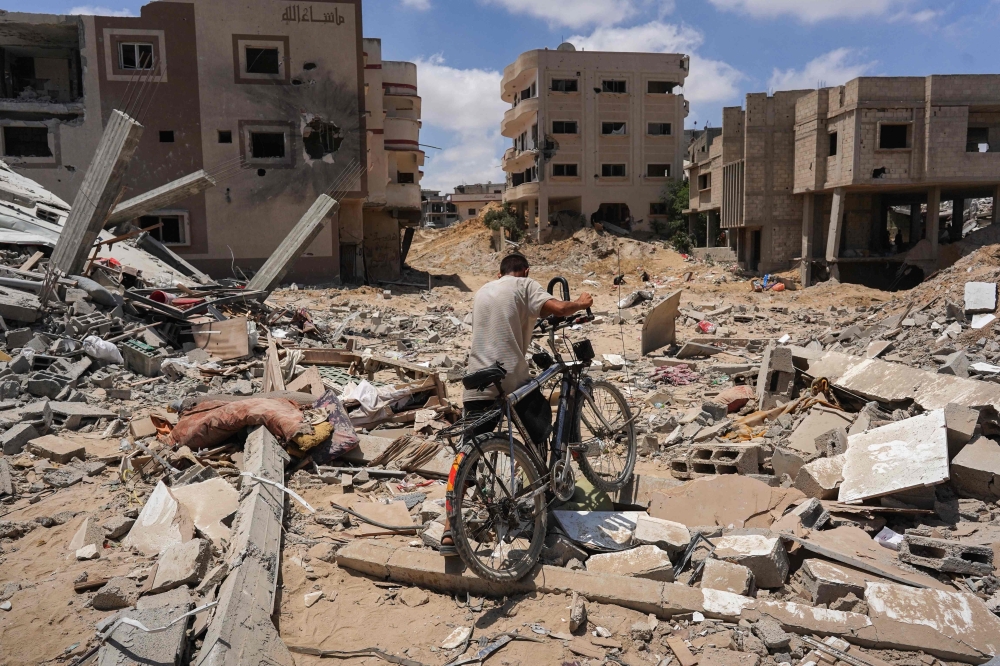 A man pushes his bicycle over rubble as displaced Palestinians return to Bani Suhayla and neighbouring towns east of Khan Yunis in the southern Gaza Strip on July 30, 2024. (Photo by Bashar Taleb / AFP)