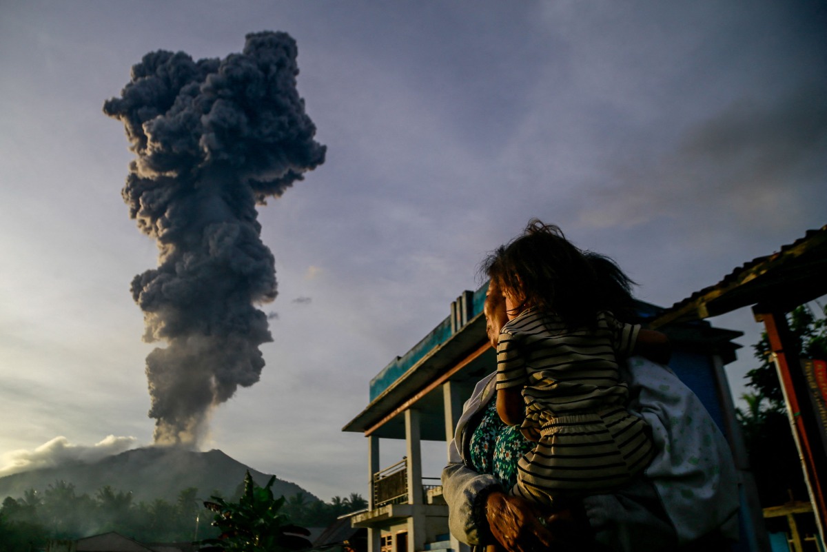 A woman and child look on at volcanic ash rising into the air during the eruption of Mount Ibu, as seen from Duono Village in West Halmahera, North Maluku province, on January 15, 2025. (Photo by AZZAM / AFP)