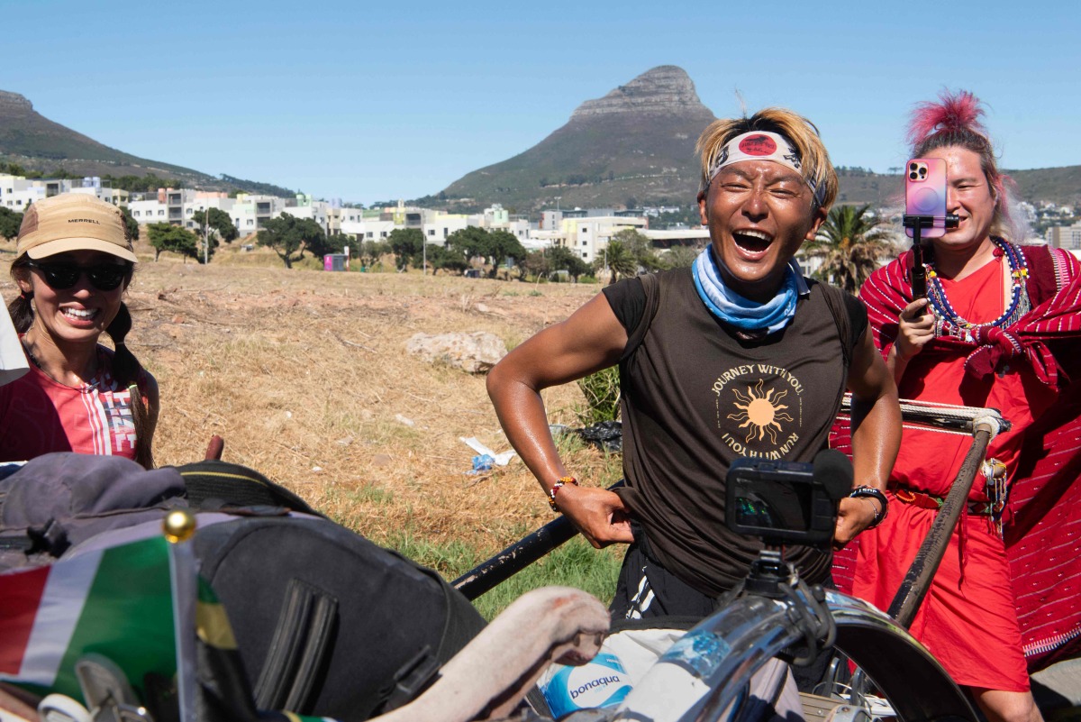 Gump Suzuki (C), the Japanese runner and social media sensation who started in July 2024 an overland journey of 6400km from Kenya to South Africa, reacts with members of his support team as he takes a rest on the side of a highway from pushing his rickshaw as he nears his end-goal at the Waterfront in Cape Town on January 26, 2025. (Photo by RODGER BOSCH / AFP)
