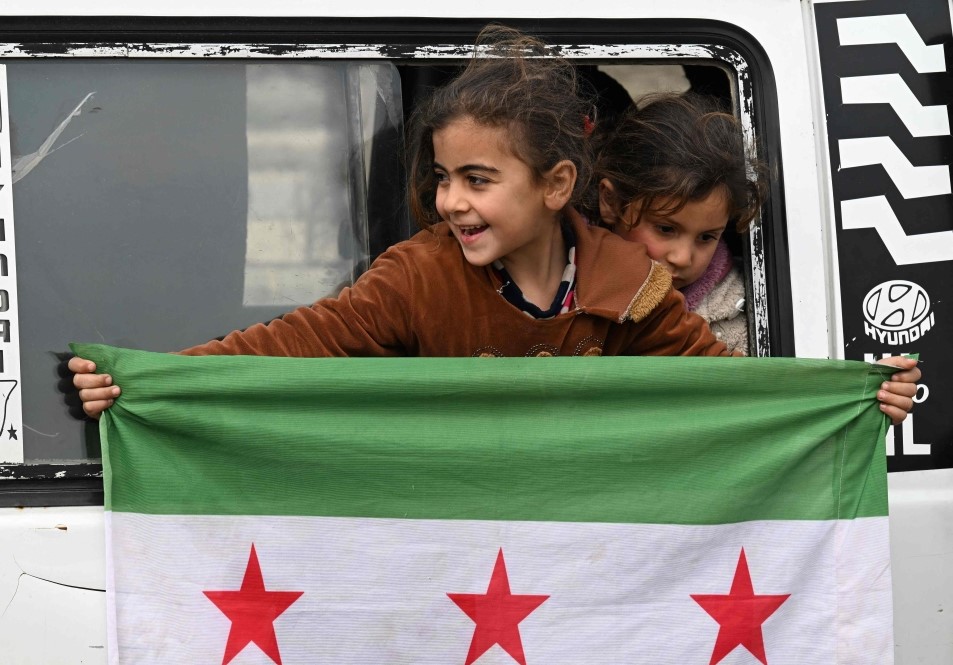 A girl holds an independence-era Syrian flag out of the window of a bus carrying displaced Syrians returning home after years of displacement in the northern Aleppo province, at the entrance of the central city of Homs on February 10, 2025. (Photo by Louai Beshara / AFP)