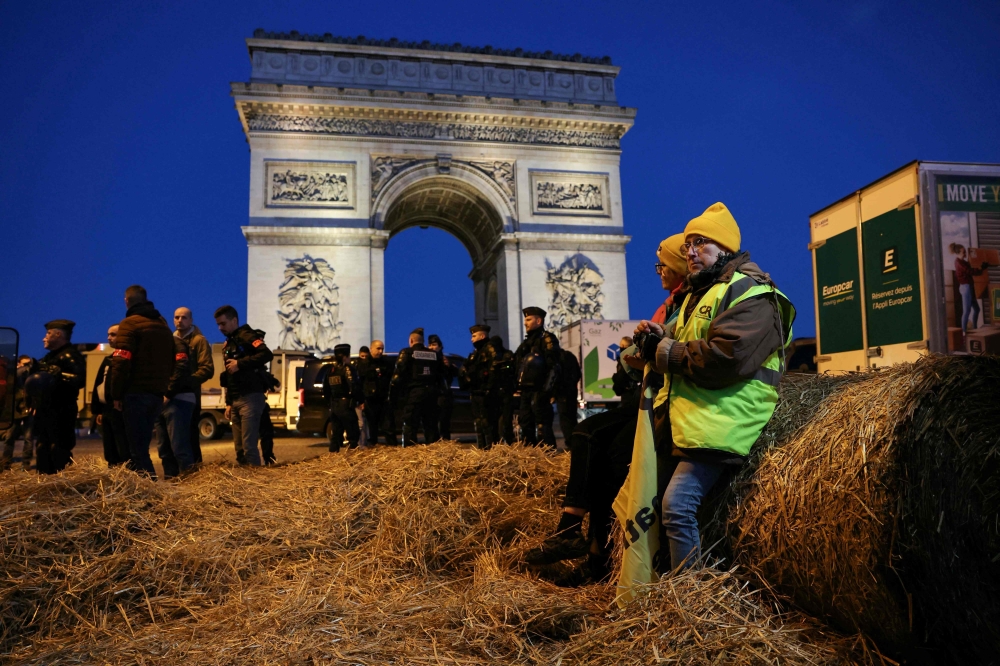 (Files) French farmers are seen sitting on a bale of hay as policemen secure the area in front of the Arc de Triomphe near the Champs-Elysees Avenue during a protest by the French farmers' union in Paris on March 1, 2024. (Photo by Thomas Samson / AFP)